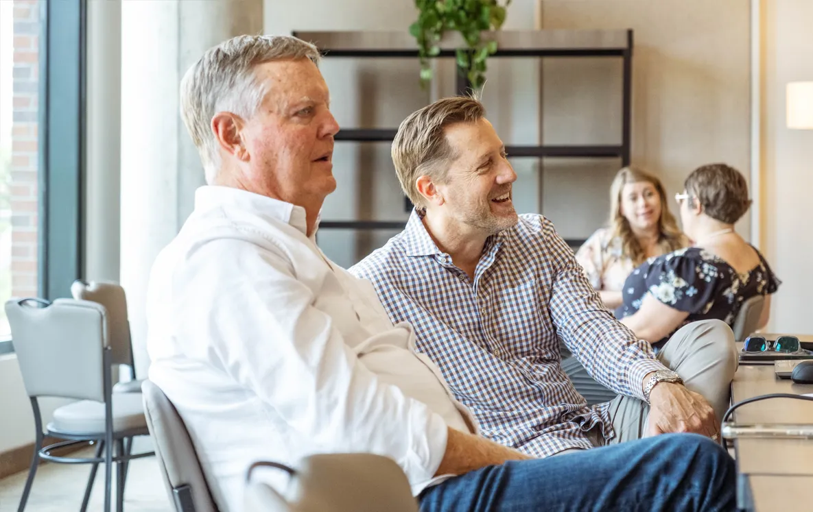 Two men, casually dressed, sitting next to one another in charis both watching something our of frame