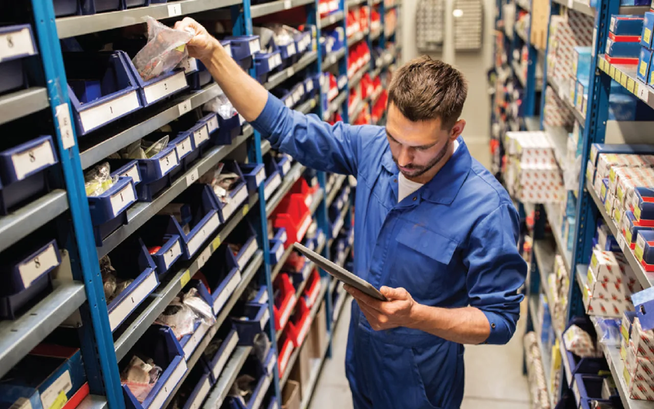 man in coveralls in a parts aisle looking at a table device and selecting an item from a parts bin